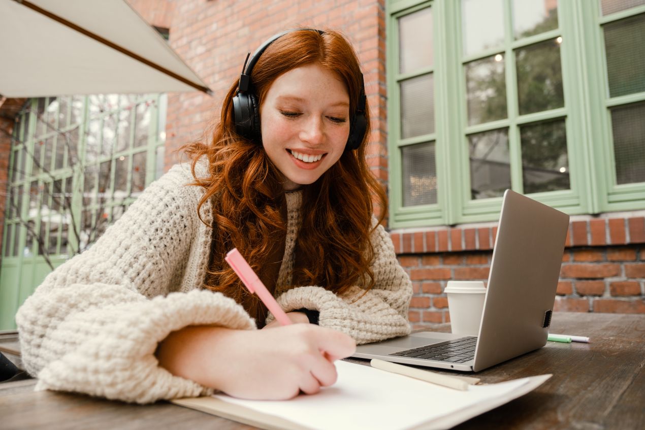 Junge Frau schreibt konzentriert mit Laptop und Kopfhoerern und kombiniert eigene Arbeit mit Inspiration zu Bachelorarbeiten schreiben lassen